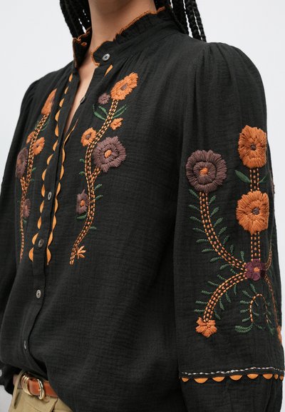 Črna majica z ročnim vezenjem cvetličnih vzorcev v oranžni in vijolični barvi. Ima zapenjanje na gumbe, nabran ramenski del in valovit rob na ovratniku.
