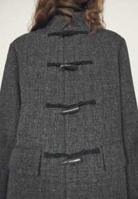 Cappotto in lana grigia con collo alto, chiusure a fibbia e due tasche frontali, caratterizzato da un tessuto strutturato e design definito.