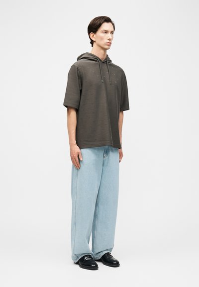 Νέος άνδρας που στέκεται, φοράει μια σκούρα πράσινη κοντομάνικη κουκούλα, ανοιχτό μπλε τζιν με φαρδιά μπατζάκια και μαύρα παπούτσια, μπροστά σε απλό γκρι φόντο.