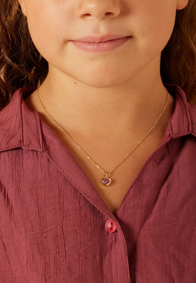 Gouden ketting met een hartvormige hanger met roze edelstenen, gedragen over een gestructureerde bordeauxblauwe blouse met een knoopdetail.