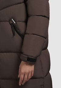 Brūns uzpūstā jaka ar quiltētām sekcijām, ar rāvējslēdzēja kabatu un melnu Velcro aproci. Gluda, spīdīga auduma tekstūra.