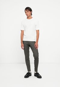 White cotton t-shirt paired with dark gray slim-fit trousers. The model wears black rounded shoes, standing against a plain white background.