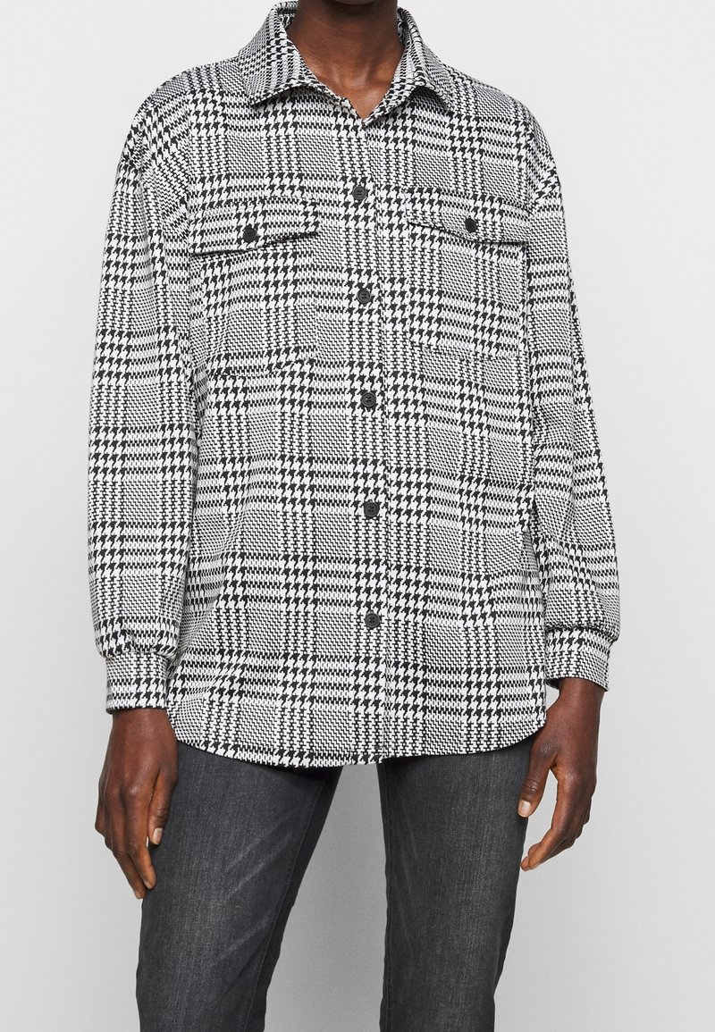 Une personne porte une chemise à boutons à motif pied-de-poule noir et blanc avec des poches poitrine et un pantalon gris foncé sur un fond uni.