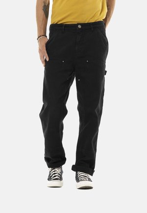 Personne portant un pantalon noir ample avec des revers roulés et des baskets noires avec des lacets blancs, debout devant un fond uni.