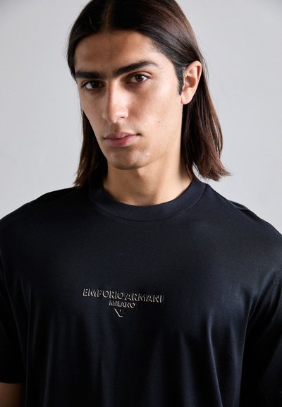 Tânăr cu părul drept, brunet, purtând un tricou negru Emporio Armani Milano, pe un fundal gri simplu.