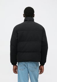 Mann, der eine schwarze gesteppte Daunenjacke und hellblaue Jeans trägt, steht mit dem Rücken zur Kamera vor einem schlichten weißen Hintergrund.