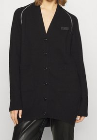 Svart cardigan med djup V-ringning, knappar och två framfickor. Materialet är stickat med ribbade detaljer vid ärmslut och nederkant.