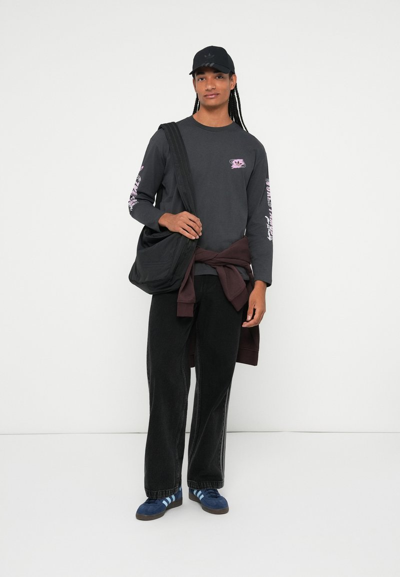 Junge Person mit langen Zöpfen, die eine schwarze Mütze, ein langärmeliges dunkles Shirt mit pinkfarbenen Grafiken, schwarze Hosen, blaue Sneaker trägt und eine schwarze Tasche dabei hat.