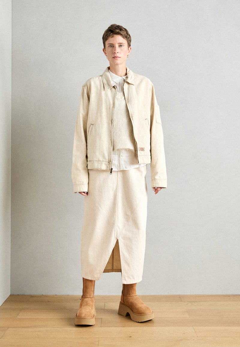 Beige Stoffjacke mit Reißverschluss und großen Taschen, über einem hellen Pullover und einem passenden Midikleid getragen; kombiniert mit braunen Plateaustiefeln.