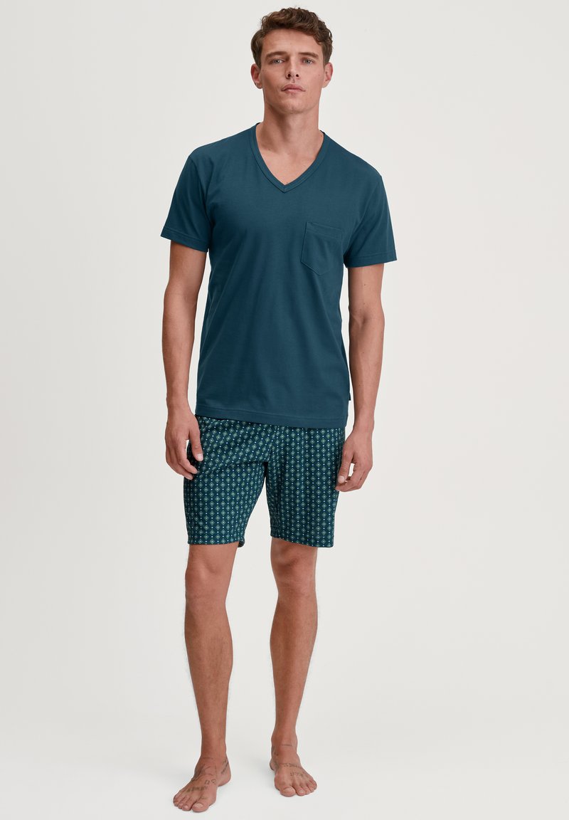 T-shirt vert canard à col en V avec une poche poitrine, assorti à un short à motifs en teal foncé et designs géométriques verts. Tissu en coton, coupe décontractée.