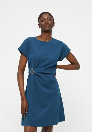 Kobieta ubrana w niebieską sukienkę z krótkim rękawem, marszczoną talią i metalową klamrą, stojąca z jedną ręką na biodrze.