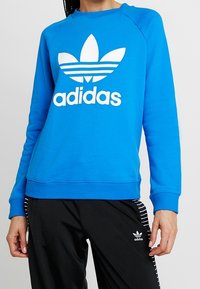 Modrá mikina s dlouhými raglánovými rukávy, bílým logem Adidas a designem trojlístku na přední straně, s žebrovanými manžetami a pasem. V kombinaci s černými kalhotami.