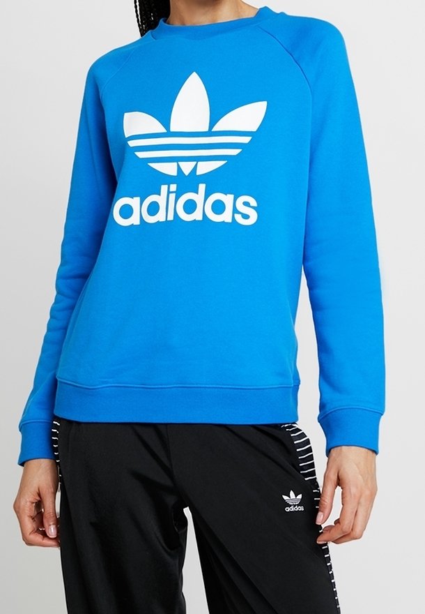 Modrá mikina s dlouhými raglánovými rukávy, bílým logem Adidas a designem trojlístku na přední straně, s žebrovanými manžetami a pasem. V kombinaci s černými kalhotami.
