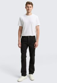 Schwarze Jeans aus Denim mit geradem Bein, kombiniert mit einem schlichten weißen T-Shirt. Das Modell trägt weiße Sneakers. Einfaches Design mit minimalen Akzenten.