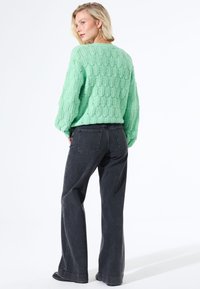 Vrouw in een mintgroene, gestructureerde gebreide trui en losse donkergrijze jeans, staande met haar handen in de zakken, terwijl ze over haar schouder kijkt.