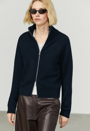 Femme blonde portant des lunettes de soleil noires, un pull zippé bleu marine, une chemise blanche et un pantalon en cuir marron foncé, debout sur un fond clair.