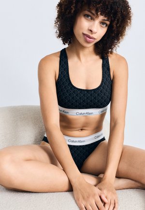 Brassière de sport noire à motifs et culottes hipster assorties avec le logo blanc de Calvin Klein sur la ceinture élastique, texture lisse, design minimaliste.