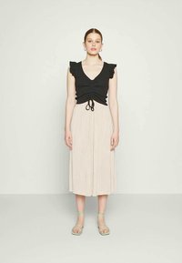 Mulher jovem vestindo uma blusa preta sem mangas com babados, saia midi plissada bege, sandálias de tirar verde-menta, em pé contra uma parede lisa.