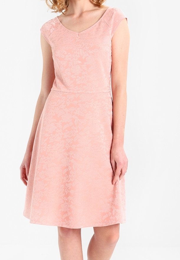 Kvinna i en knälång, kortärmad rosa klänning med subtila blommönster, stående mot en neutral vit bakgrund.