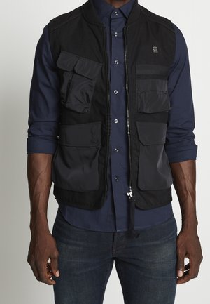 Mies, joka pukee mustan vetoketjullisen työliivin, jossa on useita taskuja, päällensä tummansinisen nappikauluspaidan ja tummat farkut.