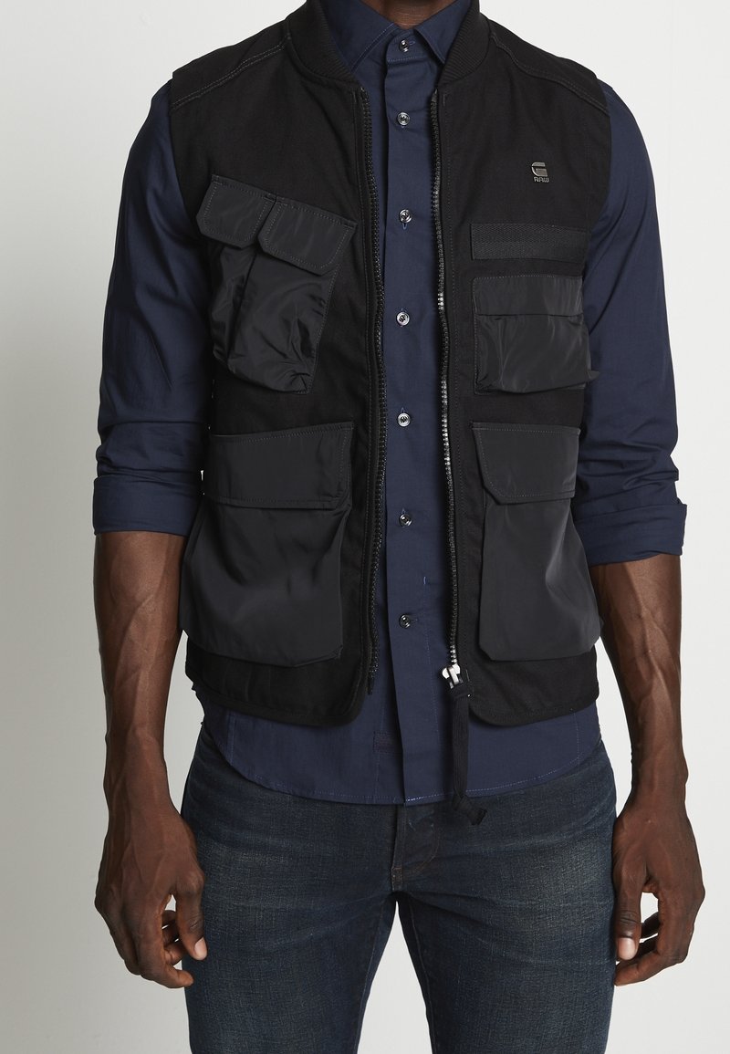 Homme portant un gilet utilitaire noir à fermeture éclair avec plusieurs poches, par-dessus une chemise bleu marine à boutons et un jean foncé.