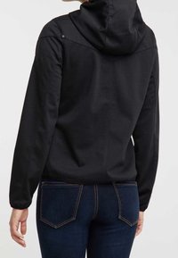 Chaqueta negra con capucha, de tela texturizada, con un cierre seguro, puños elásticos y silueta ajustada, combinada con pantalones vaqueros oscuros.