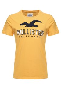 Žluté bavlněné tričko s námořnickou modrou logem Hollister a textem Kalifornie, kulatý výstřih, krátké rukávy, s hladkou texturou.