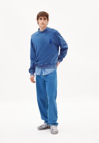 Blauer Sweatshirt mit Rundhals, auf der Brust mit einem kleinen blauen Logo versehen. Kombiniert mit einem hellblauen Denimhemd und lässigen blauen Jeans.