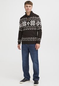 Dunkelbrauner Strickpullover mit weißen Schneeflocken- und geometrischen Mustern, mit einem Reißverschlusskragen, kombiniert mit blauen Jeans und schwarzen Sneakers.