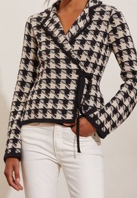 Mustapilkka ja kerma houndstooth-kietaisujakku, neulottu rakenne, lovettu kaulus, solmittava suljenta, pitkät hihat, käytetty valkoisten housujen päällä.