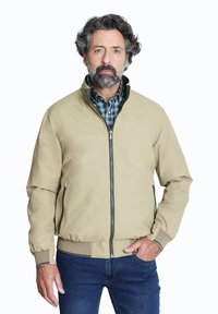 Chaqueta bomber beige con cierre frontal, puños y cuello acanalados. Llevada sobre una camisa a cuadros verde y azul, combinada con jeans azul oscuro.