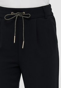 Pantalons noirs avec taille à cordon de serrage et accents métalliques, plis à l'avant et tissu lisse et texturé.