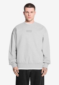 Szary sweter z okrągłym dekoltem, ściągaczami przy rękawach i prostokątną naszywką z logo na piersi. Materiał wydaje się miękki i teksturowany.