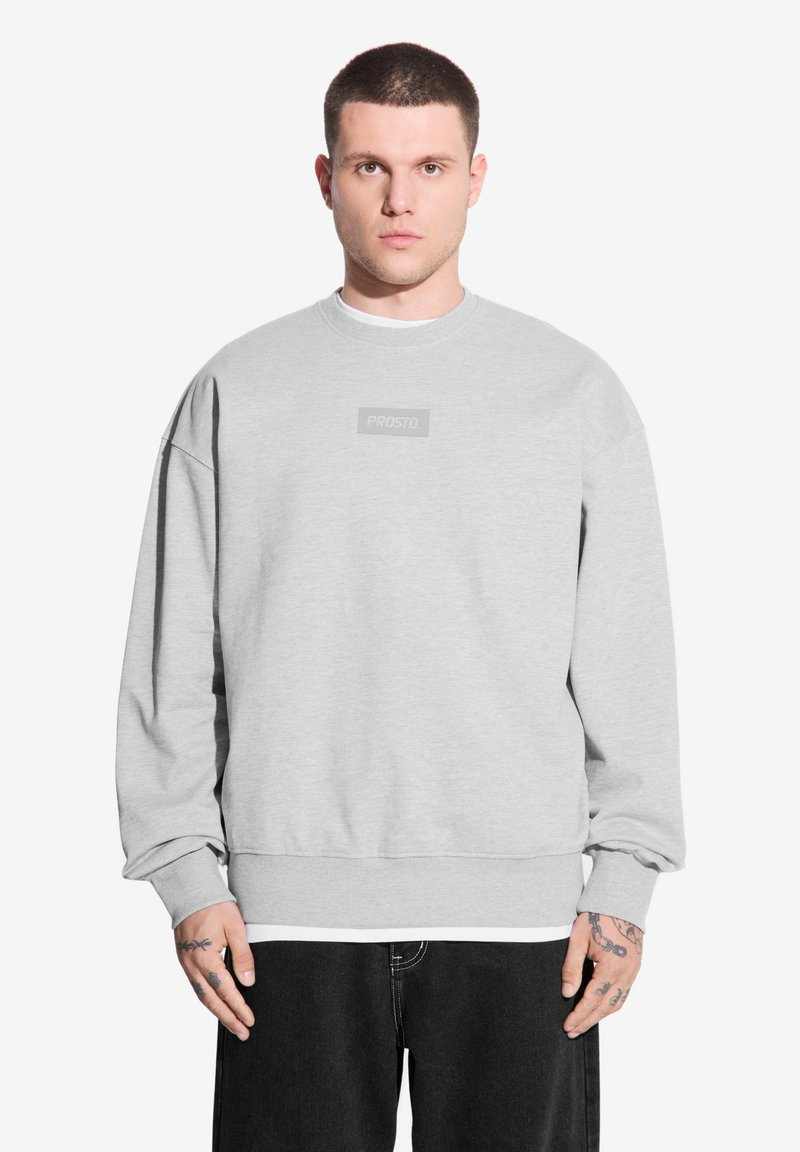 Szary sweter z okrągłym dekoltem, ściągaczami przy rękawach i prostokątną naszywką z logo na piersi. Materiał wydaje się miękki i teksturowany.