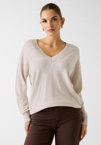 Jasnobeżowy sweter z dekoltem w serek wykonany z miękkiej tkaniny, z prążkowanym kołnierzem i mankietami; cechuje się luźnym krojem, długimi rękawami oraz minimalistycznym designem.