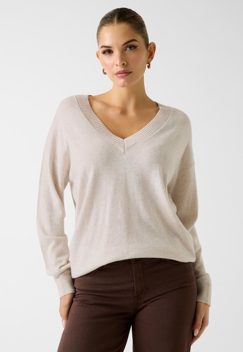 Jasnobeżowy sweter z dekoltem w serek wykonany z miękkiej tkaniny, z prążkowanym kołnierzem i mankietami; cechuje się luźnym krojem, długimi rękawami oraz minimalistycznym designem.