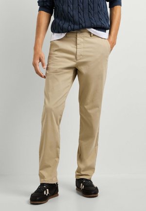 Mann trägt beige Hose, schwarze Sneaker und einen marineblauen Zopfmusterpullover mit den Händen in den Taschen vor einem schlichten Hintergrund.