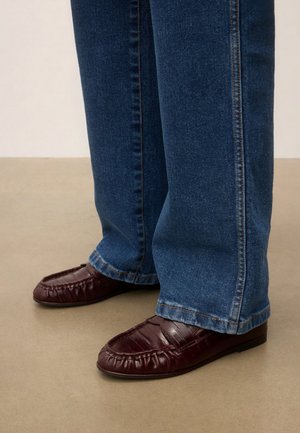 Personne portant un jean bleu foncé et des mocassins en cuir marron, debout sur un sol beige.