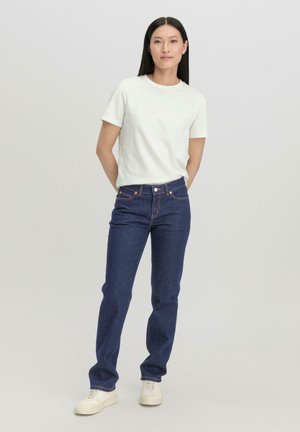 Weißes T-Shirt mit kurzen Ärmeln, dunkle blaue Jeans mit orangefarbenen Nähten und weiße Sneaker. Einfaches und klassisches Freizeitoutfit.