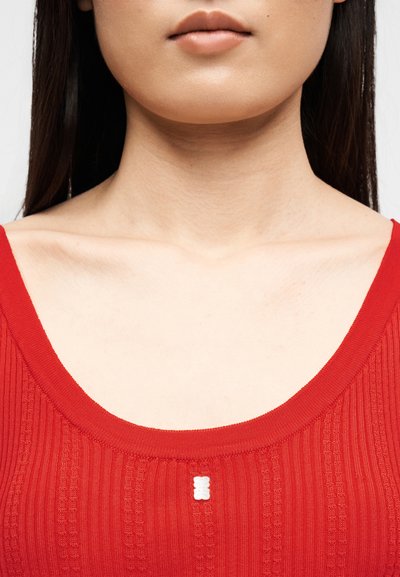 Γυναίκα που φοράει κόκκινο ριμπ μπλουζάκι με λευκή λεπτομέρεια κουμπιού στο κέντρο, φαίνονται το πηγούνι, ο λαιμός και τα κόκαλα του στήθους.