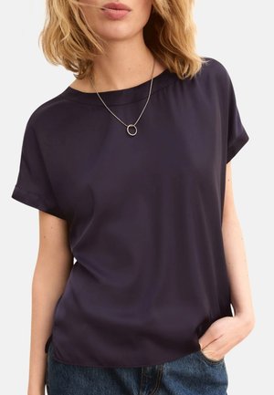 Donna con capelli biondi fino alle spalle che indossa una camicetta blu scuro a maniche corte e una collana d'argento con un ciondolo circolare.