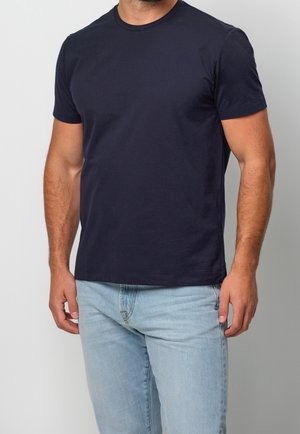 Mężczyzna ubrany w prosty ciemnogranatowy t-shirt z krótkim rękawem i jasnoniebieskie dżinsy, stojący na tle jednolitego jasnoszarego tła.