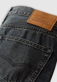 Tamsiai mėlynos džinsinio audinio kelnės su odiniu etikete, kurioje išspaustas užrašas "Levi Strauss & Co." Kelnės turi standartines kišenes ir kontrastingas siūles.