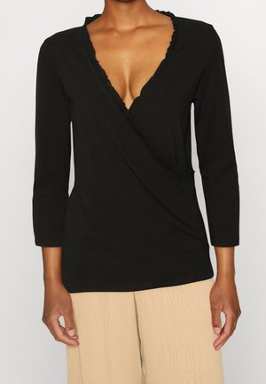 Femme portant un haut cache-cœur noir à manches longues avec un col en V à volants et un pantalon plissé beige, debout devant un fond uni.