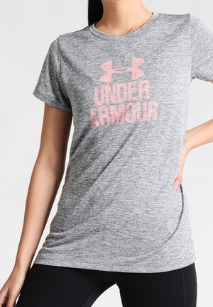 Frau trägt ein hellgraues Under Armour T-Shirt mit pinkem Logo und Text, kombiniert mit schwarzen Leggings, steht mit einer Hand in der Hüfte.