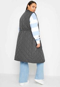 Gilet lungo grigio trapuntato con vita cinte, caratterizzato da un colletto alto e tasche laterali, indossato sopra un maglione a righe blu e bianche.