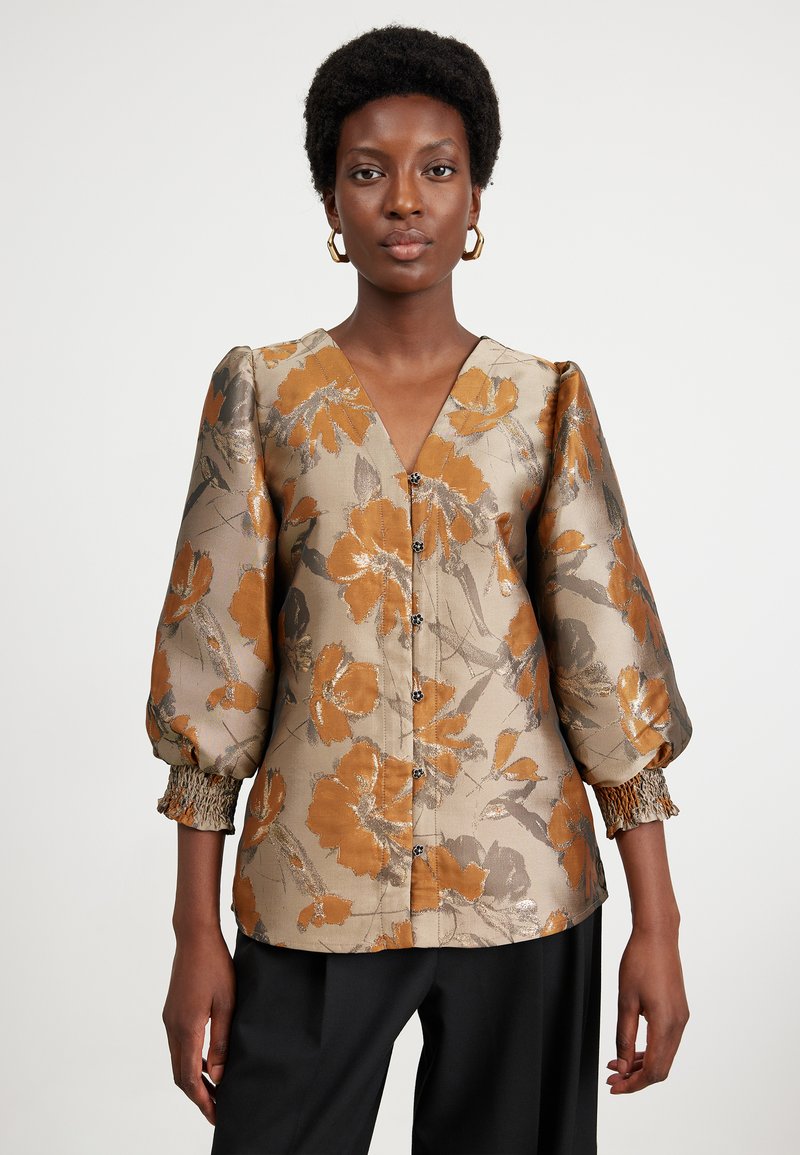 Chemisier floral avec des manches bouffantes, fleurs dorées-orange sur un fond beige. Décolleté en V et fermeture à boutons, poignets élastiques texturés.