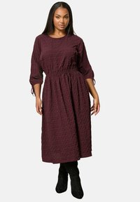 Woman with long dark hair wearing a burgundy checked midi dress with 3/4 sleeves and black knee-high boots, standing against a white background.