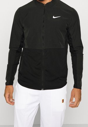 Mann trägt eine schwarze Nike-Jacke mit Reißverschluss und weißem Logo sowie weiße Hose mit kleinem mehrfarbigem Aufnäher am linken Oberschenkel.