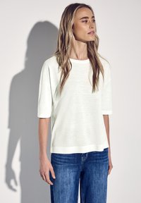 Hvid, løs pasform t-shirt med korte ærmer, lavet af et let materiale; har en rund halsudskæring og let tekstur. Bæres med blå jeans.